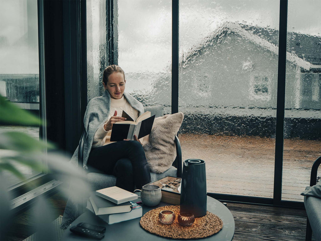 Person leser en bok i hjørne i utestuen. Det regner kraftig mot skyveglasset. Telys på bordet lager en koselig stemning.