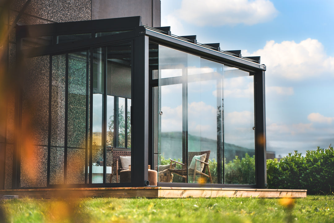 Crystal utestue på terrasse inntil steinvegg, med vintage brune møbler og planer. Utestuen er i fargen antrasitt grå, og himmelen er blå med skyer.